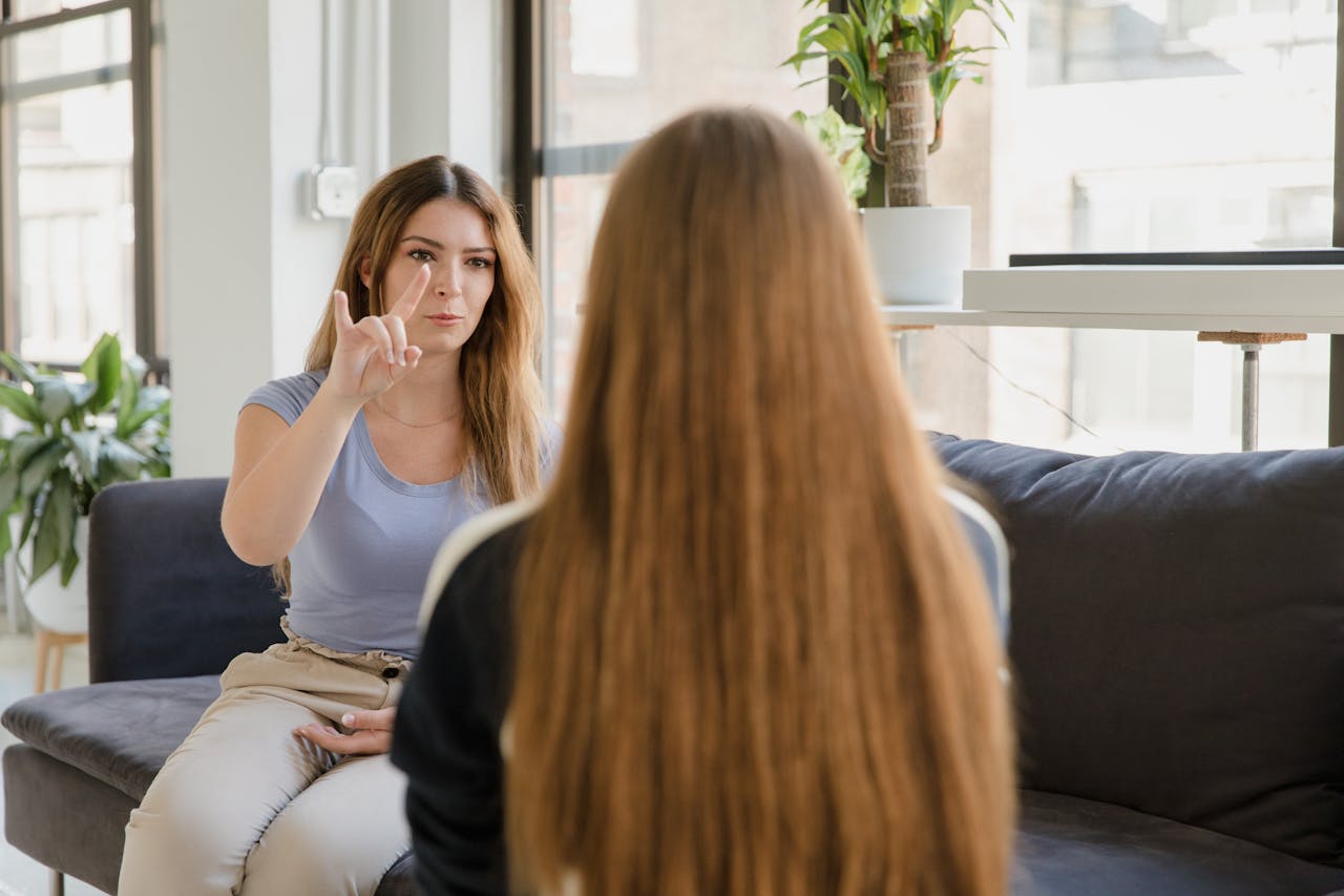 Two women engaging in sign language conversation on a couch indoors.